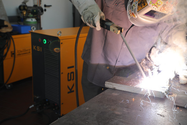 Schweißer beim manuellen Elektrodenschweißen eines Metallwerkstücks. Im Hintergrund steht eine gelbe KSI-Inverterstromquelle von Kjellberg Finsterwalde.